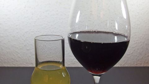 Ein großer Weinglas mit Rotwein steht neben einem kleinen Glas mit gelblichem Getränk, auf einem dunklen Tisch.