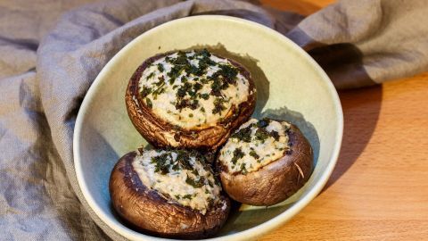 Die Pilzköpfe werden ohne Stiel in eine ofenfeste Form gelegt, Lamellen nach oben, mit Zitrone beträufelt. Ca. 20 Minuten werden die Champignons im Ofen gebacken.  Gefüllte Champignonköpfe liegen in einer Schüssel auf einem Tisch, umgeben von einem grauen Tuch. Sie wirken frisch und lecker.