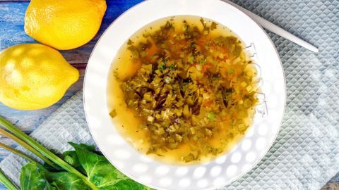 Eine Schüssel mit süßer Sauerampfersuppe steht auf einem Holztisch, umgeben von Zitronen und frischen Kräutern.