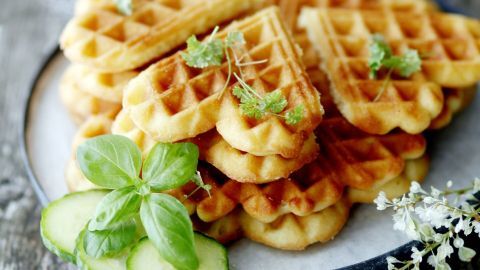 Käsewaffeln liegen auf einem weißen Teller, umgeben von frischem Basilikum und Gurkenscheiben in einer rustikalen Umgebung.