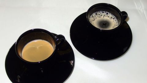 Zwei schwarze Tassen mit verschiedenen Kaffeesorten stehen nebeneinander auf einem weißen Tisch in einer stilvollen Umgebung.