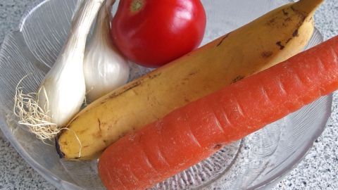 Ein Teller enthält eine Tomate, eine Banane, eine Möhre und eine Knoblauchzehe, die frisch und bunt angeordnet sind.