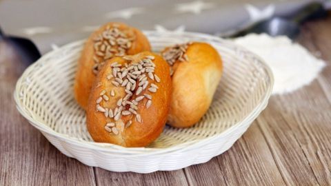 Frische, goldene Sonntagsbrötchen mit Sonnenblumenkernen liegen in einem geflochtenen Korb auf einem Holztisch.