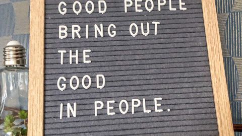 Ein Holzschild steht auf einem Tisch und zeigt den Text „GOOD PEOPLE BRING OUT THE GOOD IN PEOPLE.“ in einer gemütlichen Umgebung.