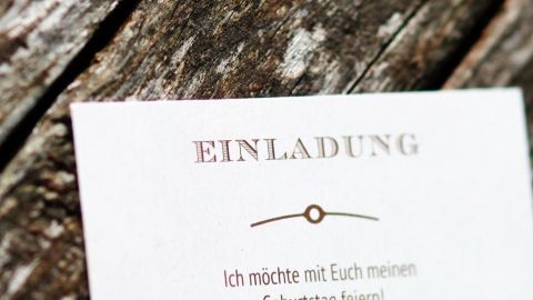 Eine Einladung liegt auf einem Holzuntergrund und kündigt den Geburtstag an, um den gemeinsam gefeiert werden soll.