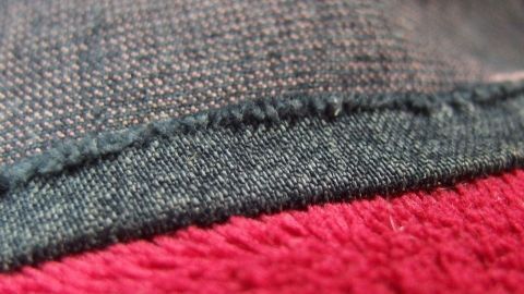 Der fokus liegt auf einem Hosen- oder Rocksaum, der sorgfältig festgeklebt wird. Ein roter und grauer Stoffschwung sind sichtbar.