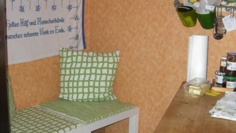 Eine weiße Küchenbank mit grün karierten Kissen steht neben einem Holztisch, vor einer orange Tapete. Text: "Gottes Hif und Menschenhände manchens schwere Ubel zu Ende."