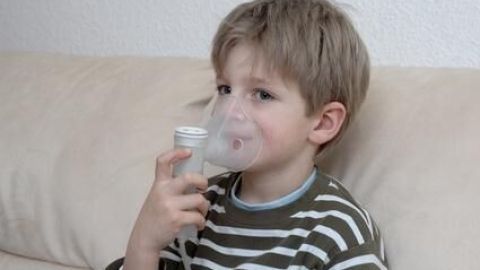 Ein Kind benutzt ein Inhalationsgerät mit Atemmaske und hält den Griff auf einem Sofa sitzend in einem gemütlichen Zimmer.