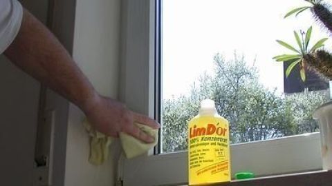 Eine Person reinigt weiße Fensterrahmen mit einem Tuch. Daneben steht eine Flasche "limDor" Grillreiniger auf der Fensterbank.