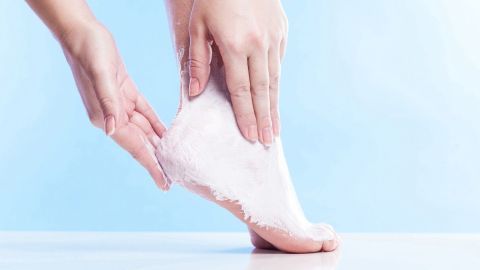 Eine Hand trägt Erkältungsbalsam auf einen Fuß auf; Hintergrund ist ein hellblauer Farbverlauf.