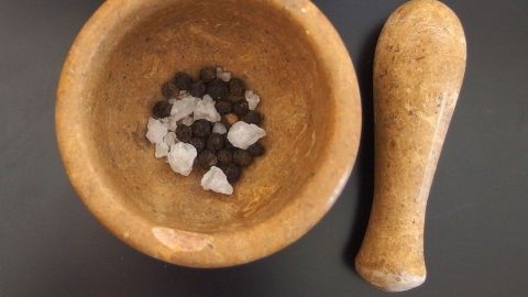 Ein Holz-Mörser enthält schwarze Pfefferkörner und grobes Salz, während der Stößel daneben liegt, bereit zum Zerkleinern.