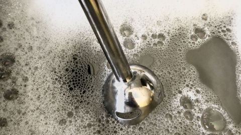 Ein Pürierstab wird im Handumdrehen sauber, wenn man ihn in das Spülwasser hält und kurz anmacht. Ein Pürierstab taucht in eine Schüssel mit seifigem Wasser und reinigt sich, um bereit für den nächsten Einsatz zu sein.