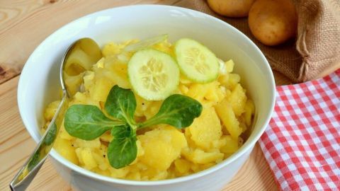 Ein frischer Schwäbischer Kartoffelsalat ist in einer weißen Schüssel angerichtet, garniert mit Gurkenscheiben und Kräutern, auf einem Holztisch.