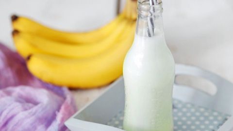 Ein Buttermilch-Bananen-Shake steht in einer Flasche auf einem Tablett, während dahinter frische Bananen liegen.