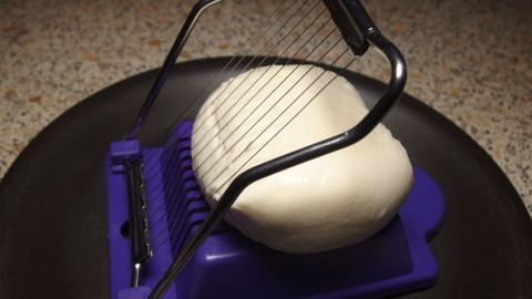 Ein Eierschneider wird verwendet, um eine runde, weiße Käse- oder Teigkugel gleichmäßig in Scheiben zu schneiden.
