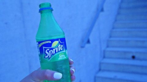 Eine Hand hält eine grüne SPRITE-Flasche, während sie in einem blauen Licht vor einer Treppe steht.