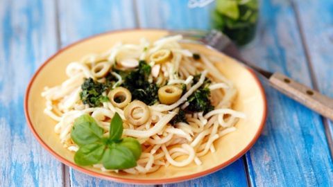 Eine Pasta mit Pesto auf einem Teller, umgeben von frischen Zutaten, steht vor einem blauen Holztisch.