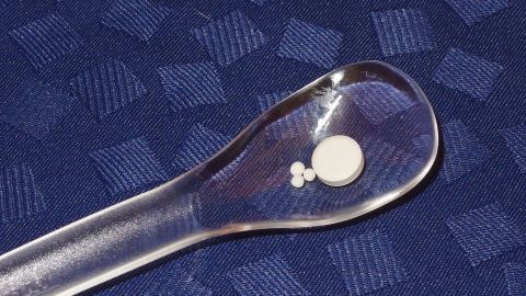 Eine Glaslöffel liegt auf einem blauen Tuch und trägt eine Tablette sowie kleine Globuli zur einfachen Verabreichung.
