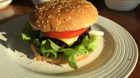 Ein saftiger Cheeseburger mit frischem Gemüse und Sesambrötchen liegt auf einem weißen Teller in einem hellen Raum.