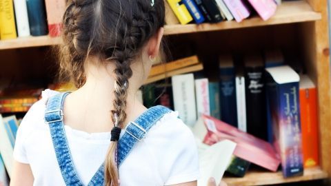 Ein Mädchen mit geflochtenem Haar steht vor einem Bücherregal und schaut konzentriert in ein Buch, um besser lesen zu lernen.