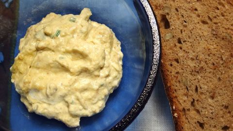 Ein cremiger, vegetarischer Brotaufstrich aus Avocado liegt auf einem blauen Teller neben einer Scheibe dunklem Brot.