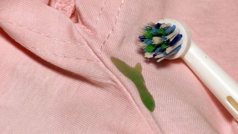 Ein grüner Fleck ist auf einem rosa Hemd zu sehen, während eine Zahnbürste zum Reinigen eingesetzt wird.