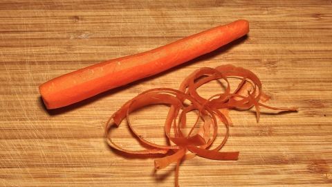 Gelbe Flecken an den Händen - nach dem Schälen von Karotten - kann man mit Speiseöl leicht entfernen. Eine orange Karotte liegt auf einem Holzbrett, daneben liegen frische Schalen, die beim Karottenschälen entstanden sind.