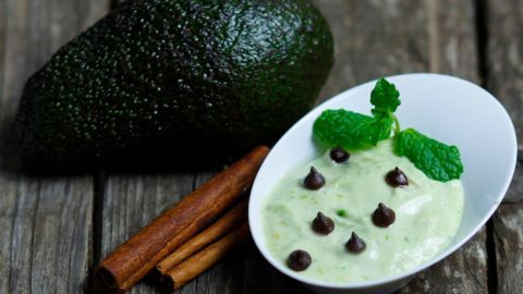 Eine grüne Avocado liegt neben einer weißen Schüssel mit cremigem Avocado-Dip, garniert mit Minze und Schokoladenstückchen.