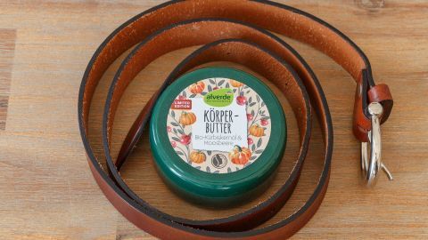 Eine grüne Dose mit der Aufschrift "Körperbutter" liegt auf einem Holztisch, umgeben von einem braunen Gürtel.