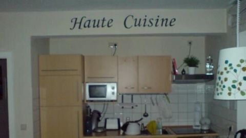 Küche mit Holzschränken, Mikrowelle und "Haute Cuisine" Wandtattoo darüber. Weißer Hintergrund, Topfpflanze auf Regal.