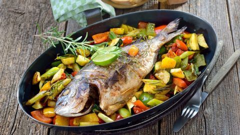 Ein gegrillter Fisch liegt auf einem Bett aus buntem Gemüse in einer schwarzen Schale, umgeben von Holzoberfläche und Brot.