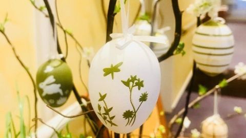 Ostereier mit Kräutermuster hängen an einem verzweigten Dekobaum, umgeben von frischen Pflanzen und Frühlingsdekoration.