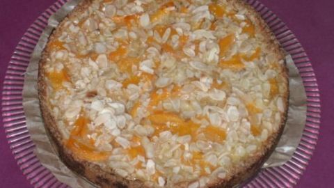 Ein runder Pfirsichkuchen auf einem Glas-Teller, belegt mit Mandelscheiben, präsentiert sich auf lila Tischdecke.