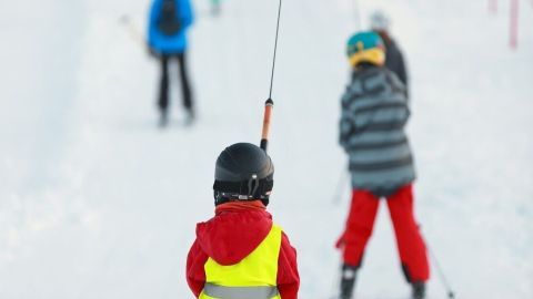 Kind mit Warnweste fährt im Skigebiet am Lift, umgeben von anderen Skifahrern auf der weißen Piste.