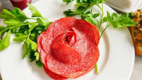 Eine kunstvoll angerichtete Salami bildet eine Blüte auf einem Teller, umgeben von frischem Grün.