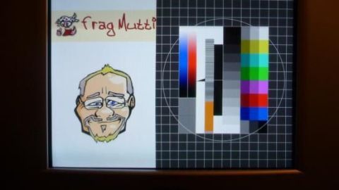 Ein Bildschirm zeigt links eine gezeichnete Männerfigur mit dem Text „Frag mich!“, rechts Farbkarten für die Kalibrierung.