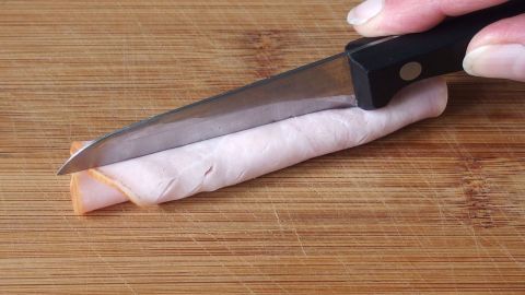 Ein Hand mit einem Messer schneidet Schinken in Würfel auf einem Holzbrett, um ihn für die Zubereitung vorzubereiten.