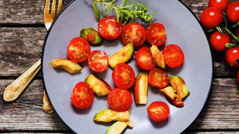 Frischer Avocado-Tomaten-Salat auf einem grauen Teller liegt auf einem rustikalen Holztisch neben einigen Kirschtomaten.