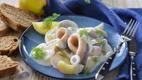 Matjesfilets in cremiger Sahnesoße liegen appetitlich auf einem blauen Teller, umgeben von Brot und frischen Äpfeln.