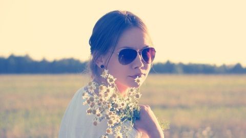 Eine Frau mit Sonnenbrille steht in einem Feld, umgeben von Blumen, und genießt die Sonne für weiche Lippen.