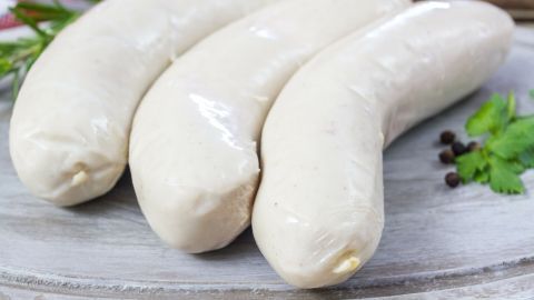Drei frische Schlesische Weißwürste liegen auf einem Holzbrett, umgeben von Kräutern, bereit zur Zubereitung.