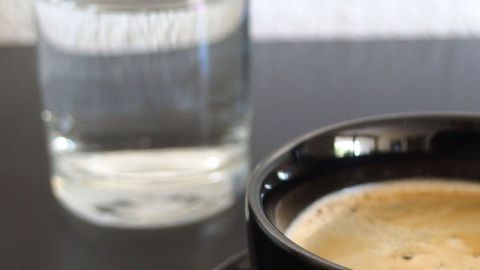 Eine schwarze Tasse mit Kaffee steht auf einem Tisch, während im Hintergrund ein Glas Mineralwasser sichtbar ist.