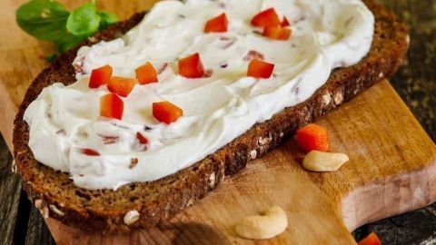 Ein Stück Brot liegt auf einem Holzbrett, belegt mit cremiger Käsecreme, dekoriert mit roten Stückchen und frischem Basilikum.