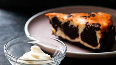 Ein saftiges Stück Kuchenteig auf einem Teller liegt neben einer Schüssel mit Mayonnaise, auf einem dunklen Tisch.