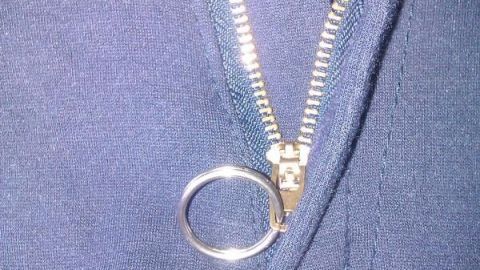 Ein simpler Trick, um deinen Reißverschluss sicher zu schließen: Fädele einen Schlüsselring durch den Zipper und befestige ihn am Knopf. Praktisch und unkompliziert für Hosen mit widerspenstigen Verschlüssen. Ein Schlüsselring sitzt an einem Reißverschluss, der gerade geschlossen wird, an einem blauen Kleidungsstück.