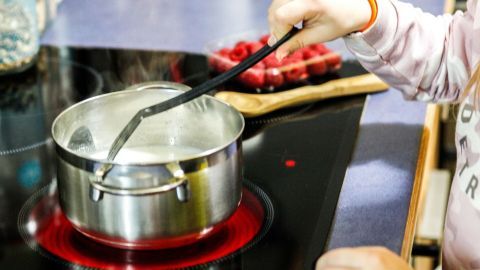 Ein Kind rührt mit einem Kochlöffel in einem Topf auf dem Herd, um ein Gericht zu kochen, während frische Zutaten im Hintergrund liegen.