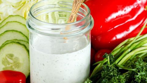 Ein Glas Knoblauch-Joghurt-Dressing steht dekorativ zwischen frischem Gemüse wie Gurken, Tomaten und Paprika.