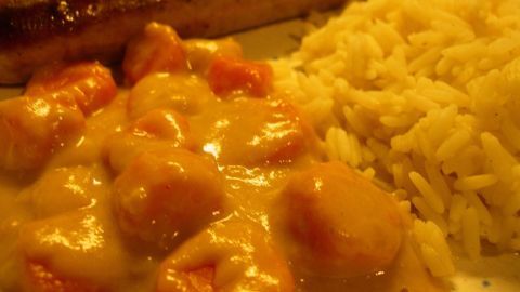 Auf dem Teller liegen Möhren mit Senfsahne, umgeben von Reis und einer Wurst, die schmackhaft angebraten ist.
