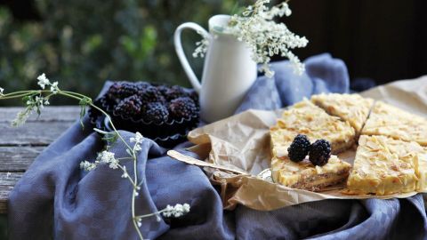 Bienenstich mit Vanillecreme ist auf einer rustikalen Holzoberfläche präsentiert, umgeben von Blumen und einer Milchkanne.