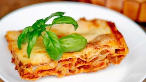 Eine appetitliche Lasagne liegt auf einem weißen Teller, garniert mit frischem Basilikum, vor einem rustikalen Hintergrund.